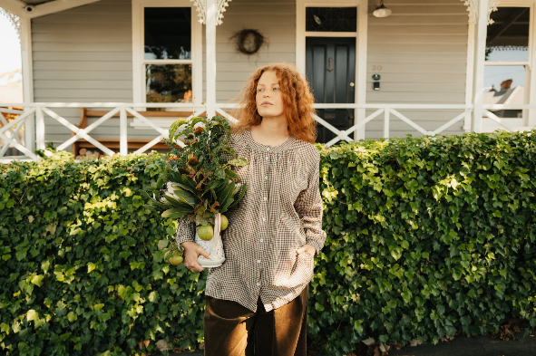 Woman holding a bag of groceries in front of a house with greenery.