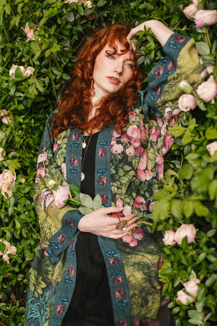 Woman wearing a floral patterned coat standing among greenery and pink flowers